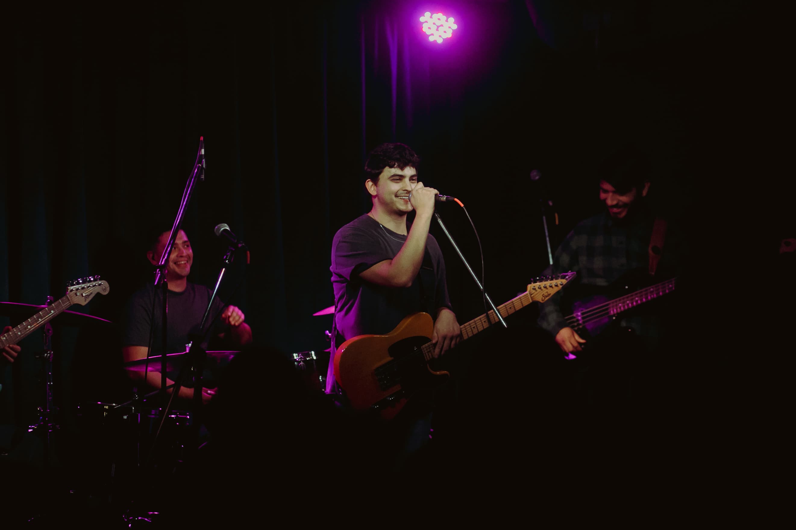 Proverbs sharing a smile on stage, with the singer at the microphone beside the drummer and bassist.