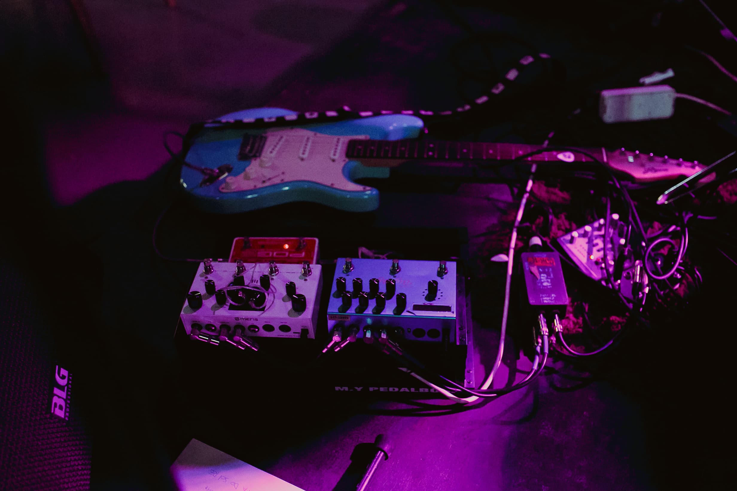 A turquoise electric guitar and effects pedals under purple stage lighting.